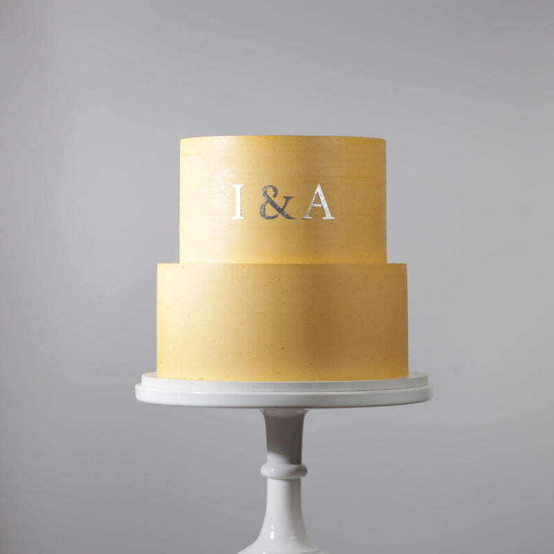 Two-tiered gold cake with 'I & A' on a gray background