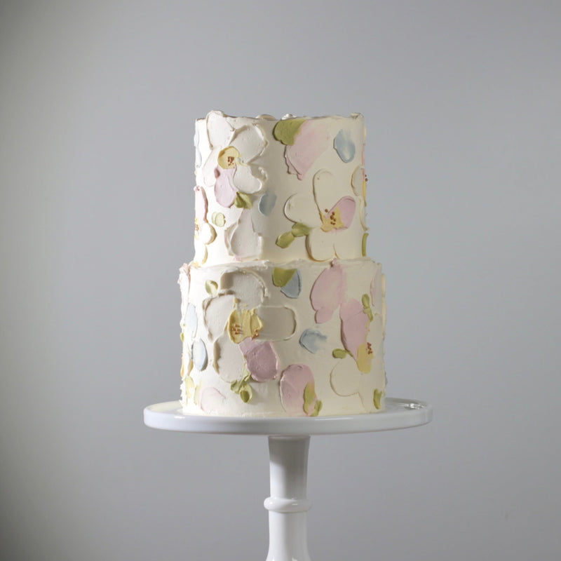 Two-tiered cake with floral patterns on a white stand against a gray background